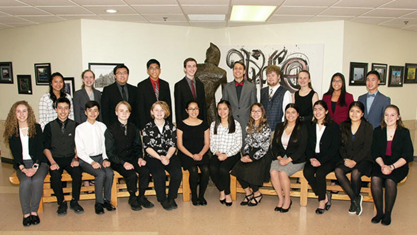 Ten students standing in the main hallway of Worthington High School with the Trojan sculpture with twelve students seated in front of them.