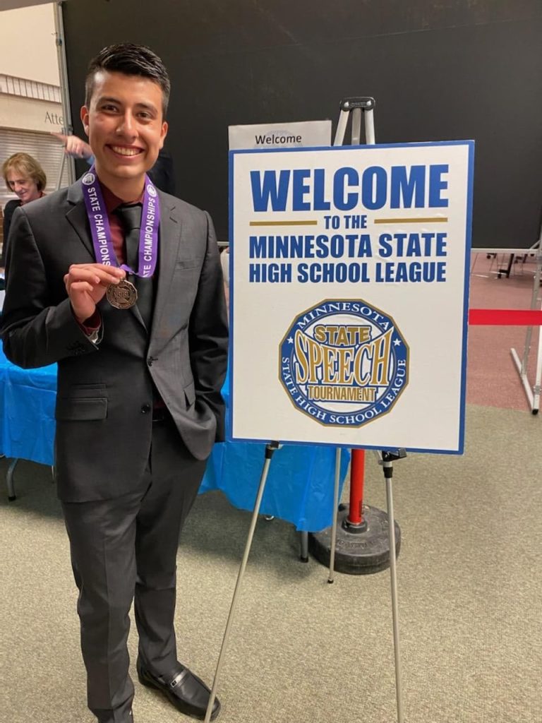 Student standing with medal next to Welcome to the Minnesota State High School League State Speech Tournament sign