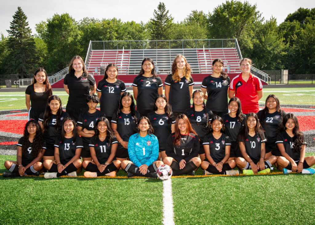 Twenty girls in 3 rows on the soccer field wearing black jerseys with 2 coaches