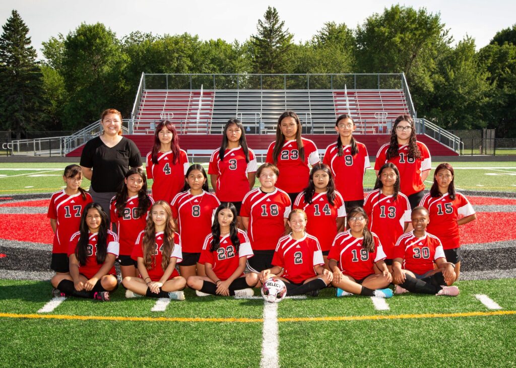 eighteen girls in 3 rows on the soccer field wearing red jerseys with a coach