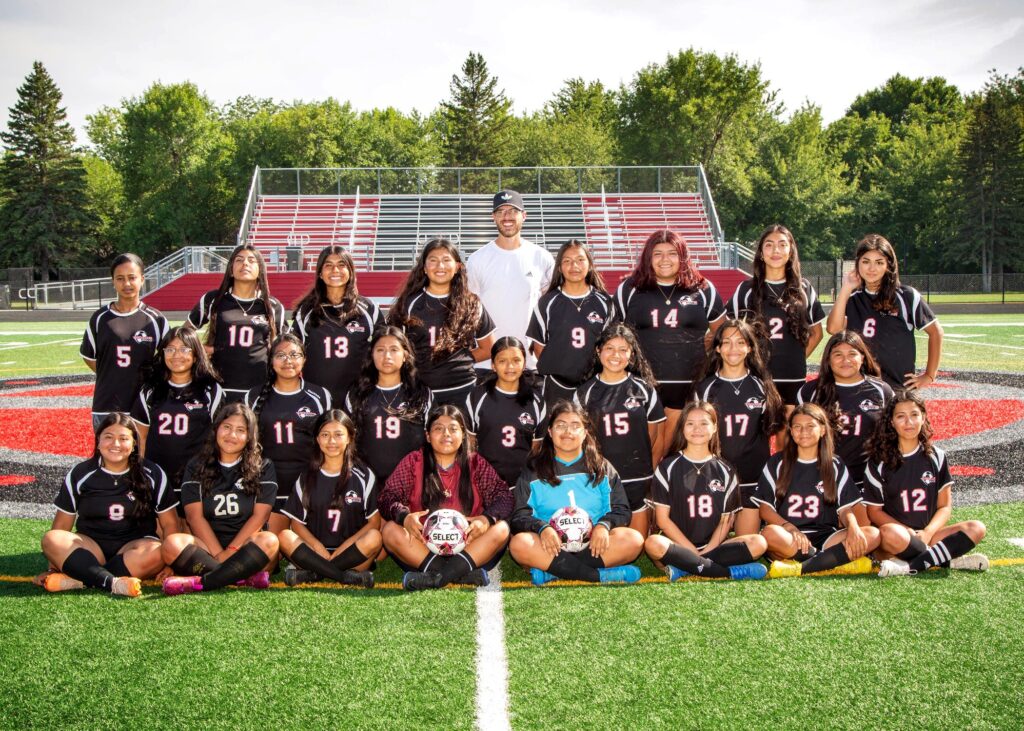 Twenty-three girls in 3 rows on the soccer field wearing black jerseys with a coach