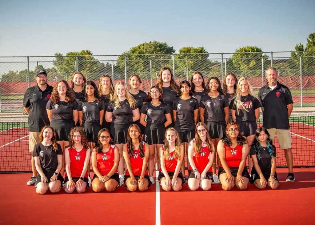 Twenty-one girls in 3 rows on the tennis court. The first row in red jerseys with the 2 standing rows in black jerseys. Coaches standing on either side.