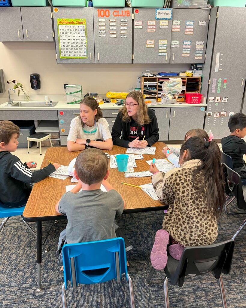 2 high school students at a table with 4 elementary students at a table