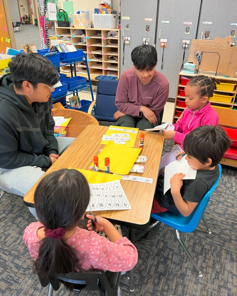 2 high school students at a table with 3 elementary students doing a math activity
